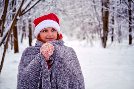 A woman is wrapped in a blanket against the background of a winter park. Red-haired woman on Christmas and New Year Eve walks in the winter in red clothesの写真素材