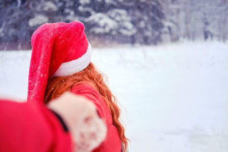 A woman leads a man into a snowy park - follow me. A couple on Christmas and New Year Eve walks outdoors in a red hat and jacket.の写真素材