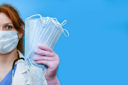 Doctor with medical mask in hand, copy space. A nurse hands out face masks on a blue background. Concept of protective medical clothing for the coronavirus pandemicの写真素材