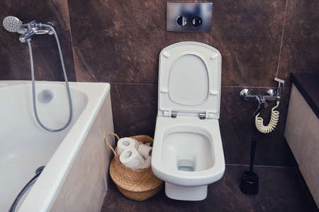 White toilet with hygienic shower next to the bathtub and washbasin cabinet. Toilet paper basket and brown stone tilesの写真素材