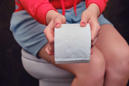Woman holds out a roll of toilet paper while sitting on the toilet, copy space for text. The concept of stomach disease, poisoning and diarrhea problemsの写真素材