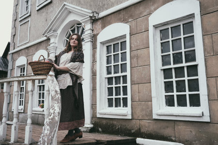 A woman with a basket in hand stands on the stairs at the railing of an old house with white windows