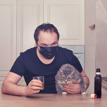 A man in a face mask drinks strong alcohol while sitting with a bottle at the kitchen table with a pet. Problems with alcoholism during the epidemic, isolation and lockdownの写真素材