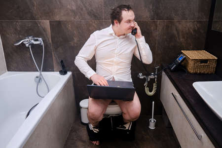 Funny man in suit with laptop and phone at home office, work without pants in the toilet. Fun internet work from home during the corona virus epidemic, conceptの写真素材