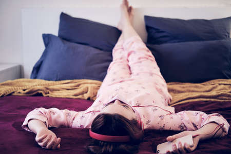 A woman in a makeshift mask is isolated during quarantine by the coronavirus. The woman on the red bed listens to music from the phone in the headphonesの写真素材
