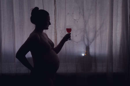 Adult pregnant woman with a glass of wine, shadow. Problems with alcohol during pregnancy, silhouette of the expectant mother at the night windowの写真素材