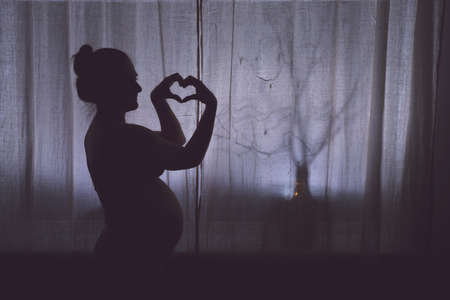 Pregnant woman shows a hand gesture sign of love on Valentine Day, silhouette against the background of a night windowの写真素材