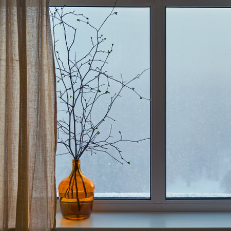 Branches of a tree with blooming leaves stand in a vase by the window against the background of a snow stormの写真素材