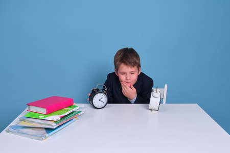 Boy schoolboy with a alarm clock in his hands at the table on a blue backgroundの写真素材