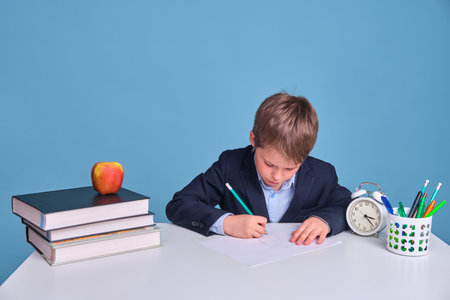 Schoolboy boy in uniform with books and a pen writes on a sheet of paper, blue backgroundの写真素材