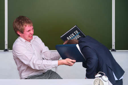 Angry teacher beats a schoolboy with a book in the classroom at the blackboard, copy spaceの写真素材