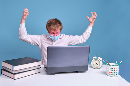 Angry man in medical face mask works behind laptop on blue studio background