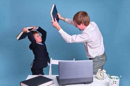 A man and a schoolboy boy fight with books on a blue background. Challenges to work and study from home during the coronavirus pandemicの写真素材