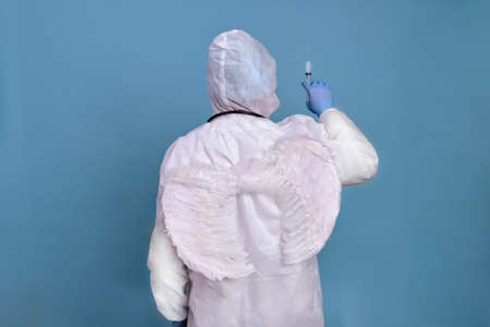 Doctor stands with his back with white angel wings and a syringe, blue backgroundの写真素材