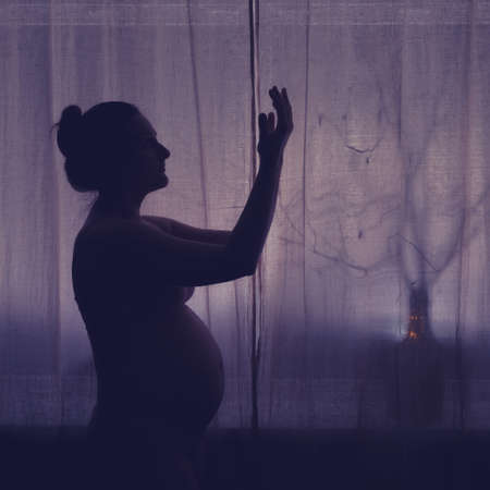Pregnant woman raised her hands up asking for help. Pregnancy problems, silhouette of the expectant mother at the night window in prayer to Godの写真素材