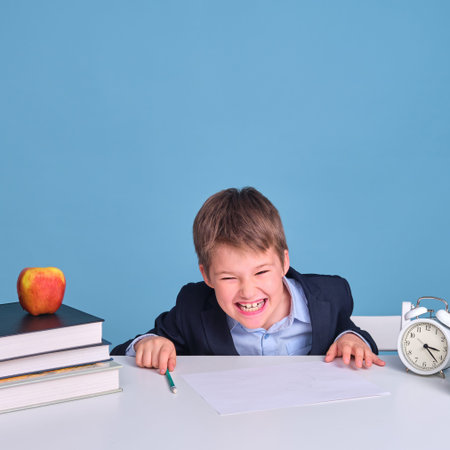 Schoolboy boy in uniform with textbooks and pen laughs while sitting at a school desk, blue...