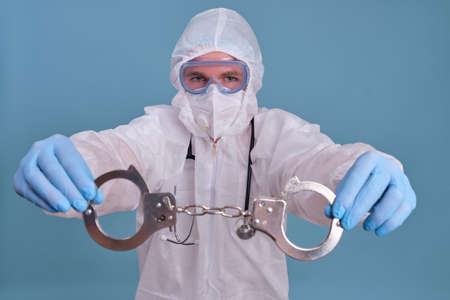 A paramedic in a protective uniform in a medical face mask with handcuffs in his handsの写真素材