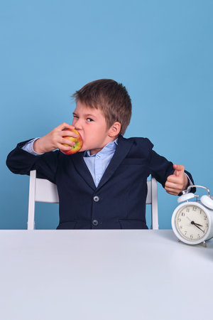 Junior in school uniform eats an apple at his desk, blue background