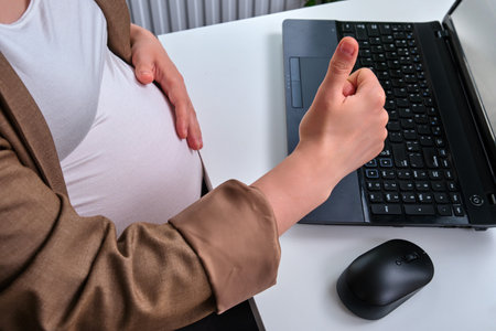 Hands of a pregnant woman with a thumb up near the computer keyboardの写真素材