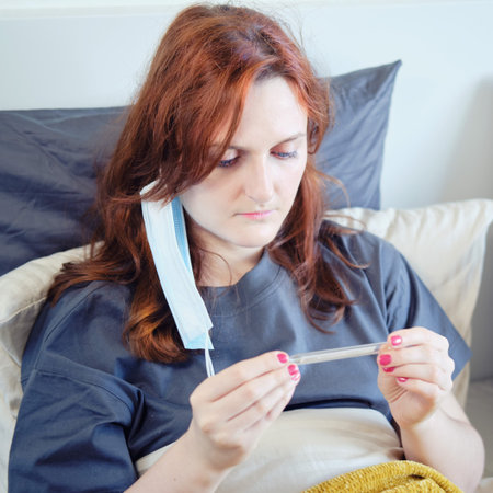 Redhead woman with coronavirus with mask on her face looks at a thermometer. woman with a flu virus in bed at home in a medical maskの写真素材