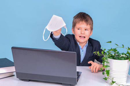 A boy in a school suit with a medical face mask at a table with a laptop, copy space on a blue...
