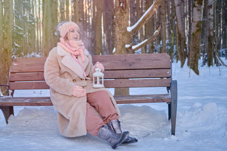 A happy woman is sitting on a bench with a lantern in her hands, a winter park with snow-covered treesの写真素材