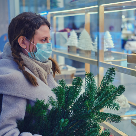 A woman in a medical face mask with a Christmas tree in her hands looks at a shop window with New Year giftsの写真素材