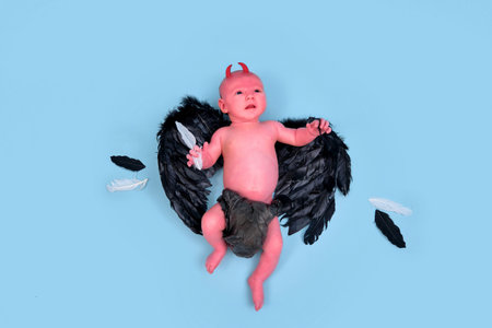A newborn baby boy with black demon wings on a blue studio background, copy space. An infant...