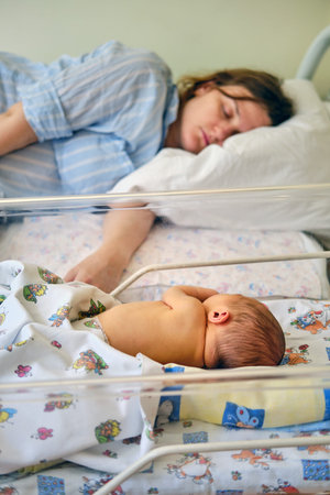 Mom is sleeping in the ward with a newborn baby, a maternity hospital bracelet on her arm. A newly born child in a clinic crib behind a transparent glassの写真素材