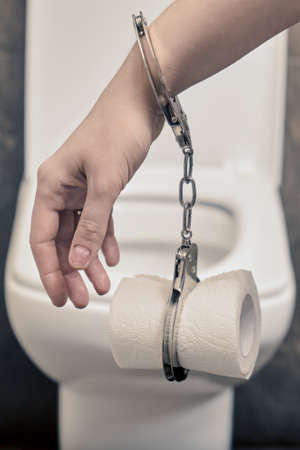 Hand of a man with toilet paper in handcuffs on the background of the toilet. Concept of constipation problems and abdominal stiffnessの写真素材