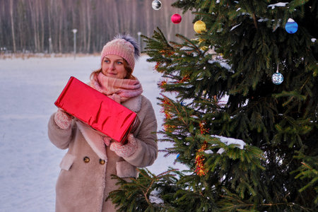 A happy woman in winter clothes stands with a gift at a Christmas tree in a snowy forest on New Year...