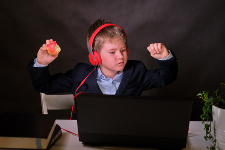 A happy boy in a school suit with headphones listens to music at the computer, copy space on a dark studio backgroundの写真素材