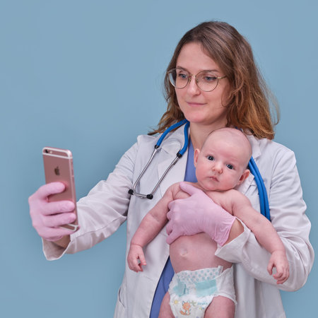 Pediatrician doctor holding iPhone Apple and newborn baby, blue studio background. Happy nurse in...