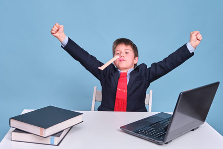 A boy in a school suit raised his hands in a victorious gesture at the computer during distance learning, copy space on a blue studio backgroundの写真素材
