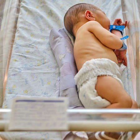 A newborn baby with a maternity hospital bracelet on his arm is sleeping in a crib. A newly born child in a clinic bed behind a transparent glassの写真素材