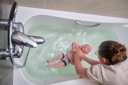 Mom and son are engaged in infant swimming in the home bathroom. A woman helps a newborn baby to...