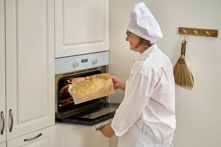 Pie in a baking dish is placed in a preheated oven. An adult woman in white chef clothes cooking pie in a beige kitchenの写真素材