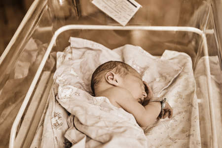 A newborn baby with a maternity hospital bracelet on his arm is sleeping in a crib. A newly born child in a clinic bed behind a transparent glassの写真素材