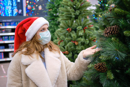 A woman in a medical face mask chooses a faux Christmas tree in a store with New Year giftsの写真素材