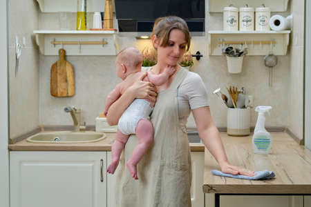 A woman holds an infant baby in her arms while cleaning in a home kitchen, problems with household chores after the birth of a childの写真素材