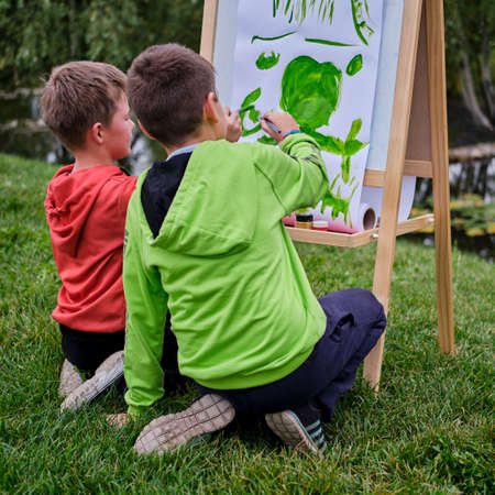 Children learn to paint on canvas, easel with paper. Boy artist paints nature and trees by the water surface of the riverの写真素材