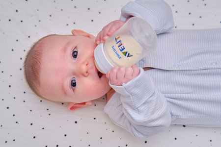 Happy baby is smiling on her own holding a bottle Philips Avent in her hands. Three month old child keeps the mixture himself lying in bed- Moscow, Russia, October 05, 2021のeditorial素材