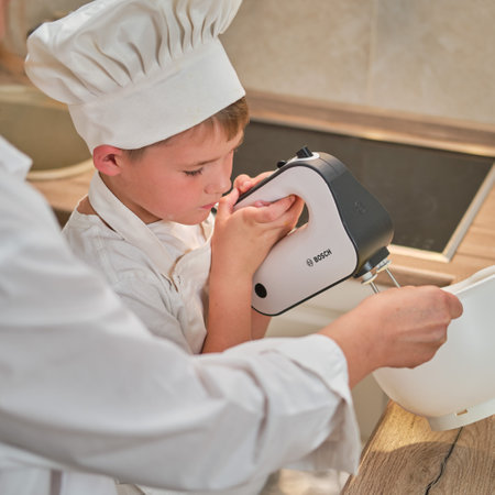 Boy in white chef clothes beats eggs with a Bosch mixer to make a pie in his home kitchen - Moscow, Russia, September 2, 2021のeditorial素材