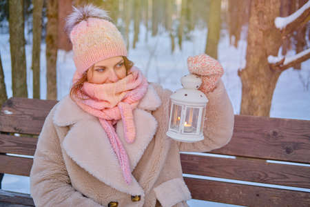 A happy woman is sitting on a bench with a lantern in her hands, a winter park with snow-covered treesの写真素材