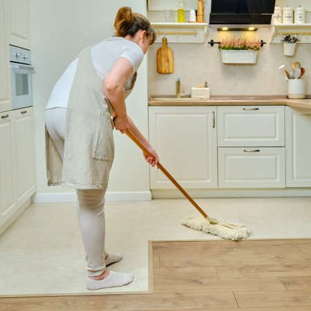 A woman in an apron cleans the floor with a mop in a home kitchenの写真素材