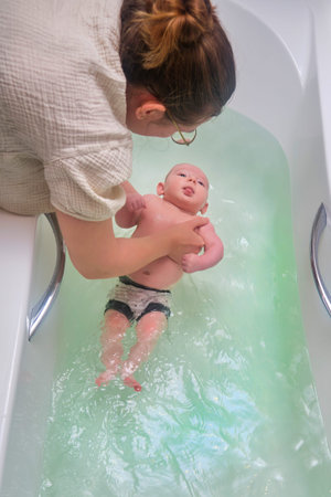 Mom and son are engaged in infant swimming in the home bathroom. A woman helps a newborn baby to...