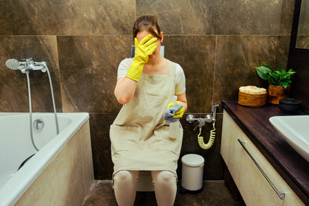 Emotions of shame in a woman sitting on the toilet while cleaning the bathroom. Disinfection with yellow rubber gloves in the washroomの写真素材