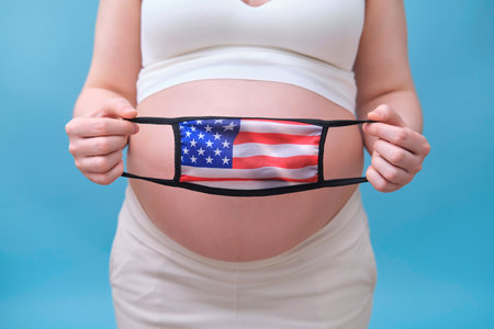 Medical face mask with the US flag in the hands of a pregnant woman, studio shot on a blue backgroundの写真素材