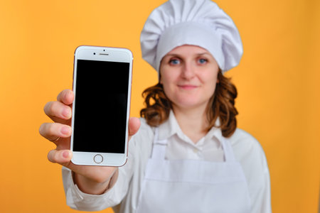 Happy woman chef showing iphone screen on yellow studio background, mockup copy space - Moscow, Russia, October 20, 2021のeditorial素材
