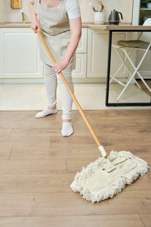 A woman in an apron cleans the floor with a mop in a home kitchenの写真素材
